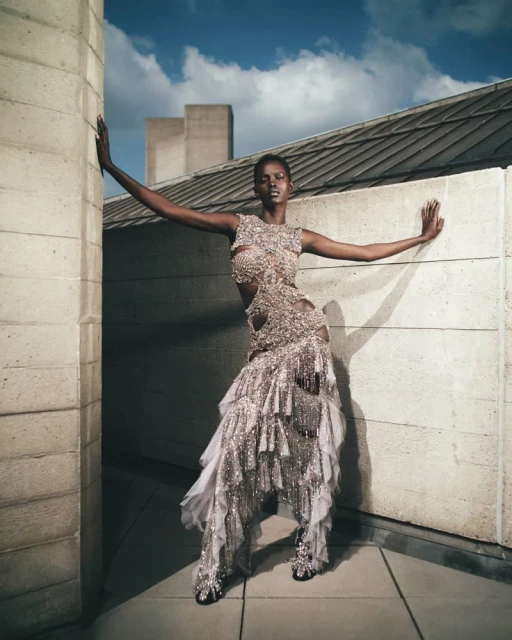 Fashion shot of an African female adult model in an ornate cut-out dress, posing against a concrete wall under a partly cloudy sky outdoors.
