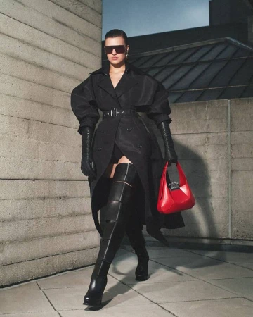 Model wearing a structured black coat, thigh-high boots, and dark sunglasses holding a red handbag against an architectural concrete wall.