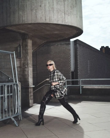 Model in black sunglasses wearing a patterned coat and tall black boots outdoors on a concrete rooftop.