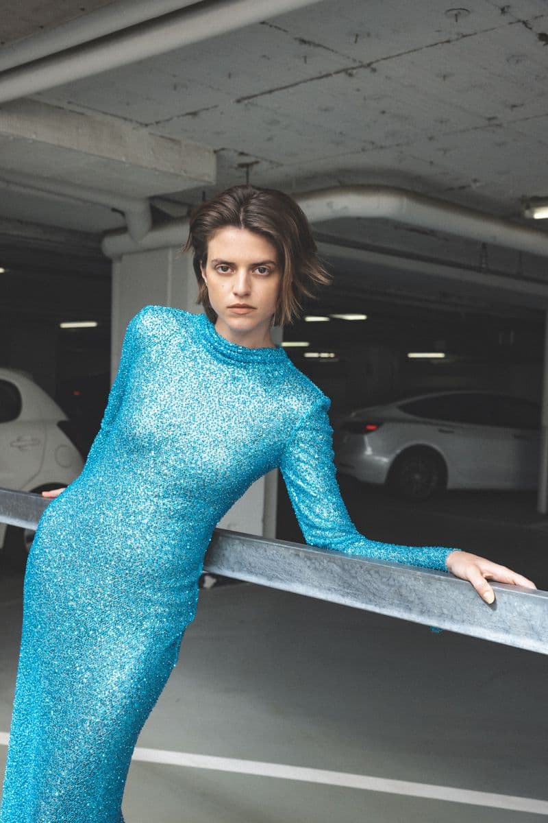 Elegant woman in a sparkly turquoise dress leans against a railing in a parking garage, showcasing a sophisticated and modern fashion statement.