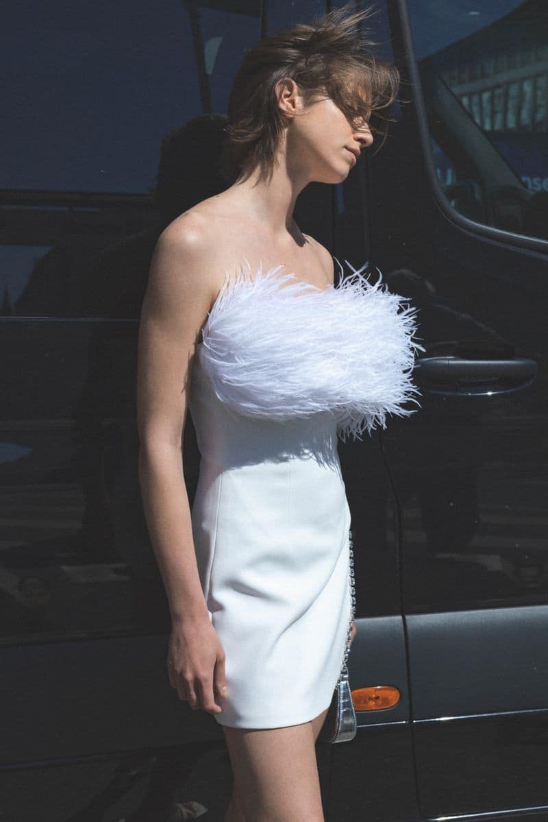 Elegant woman in a short white dress adorned with feathers, posing outdoors with natural light and a blurred vehicle background.