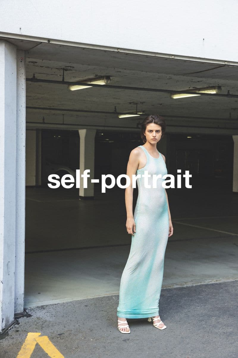 Woman in long dress poses in parking garage. Full-length shot, eye-level angle, natural light, fashion editorial style. White high-heeled sandals.