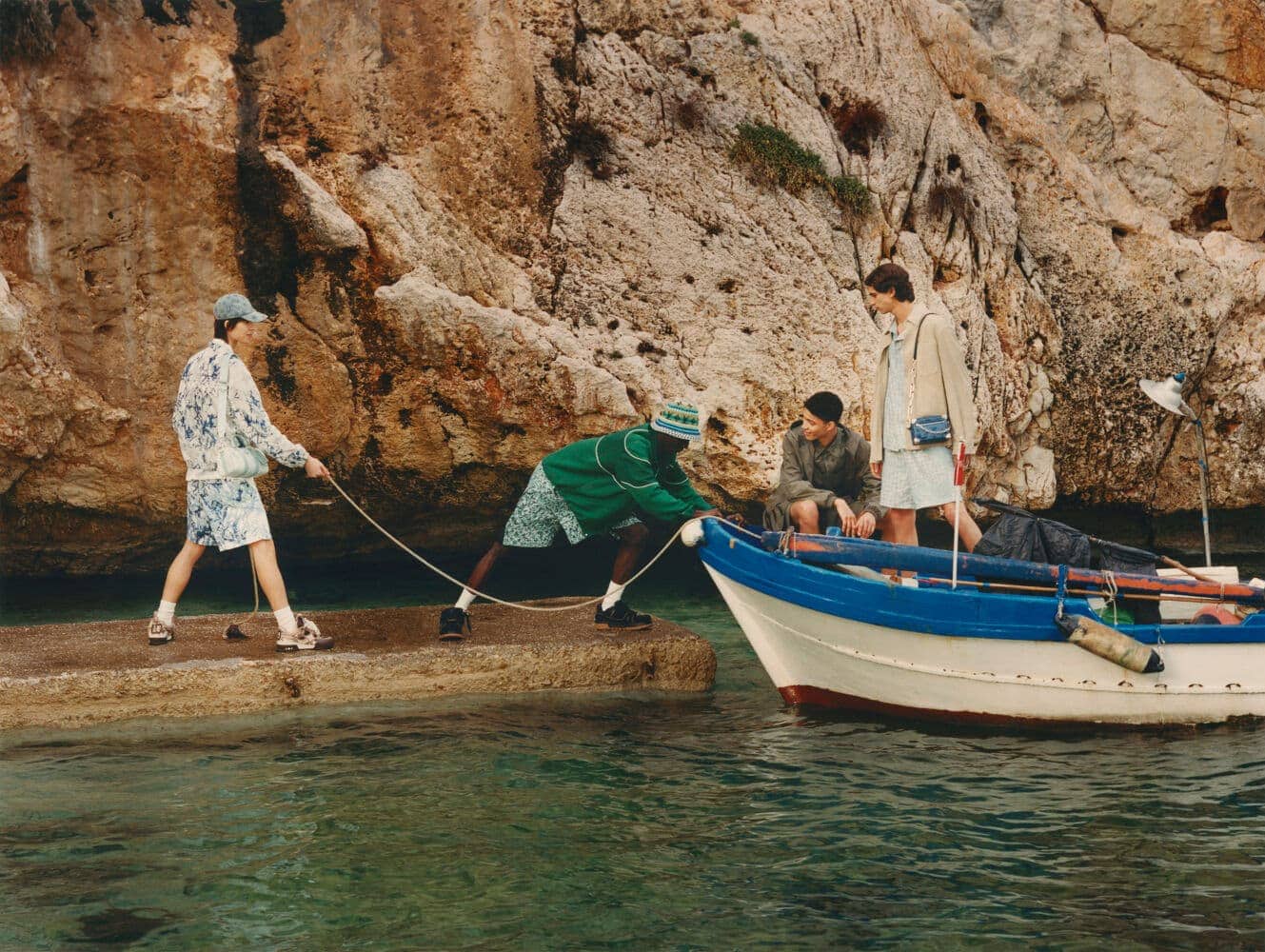 Four people and a boat at the water's edge, featuring two men pulling a rope attached to a small boat, with a rocky coastline backdrop.