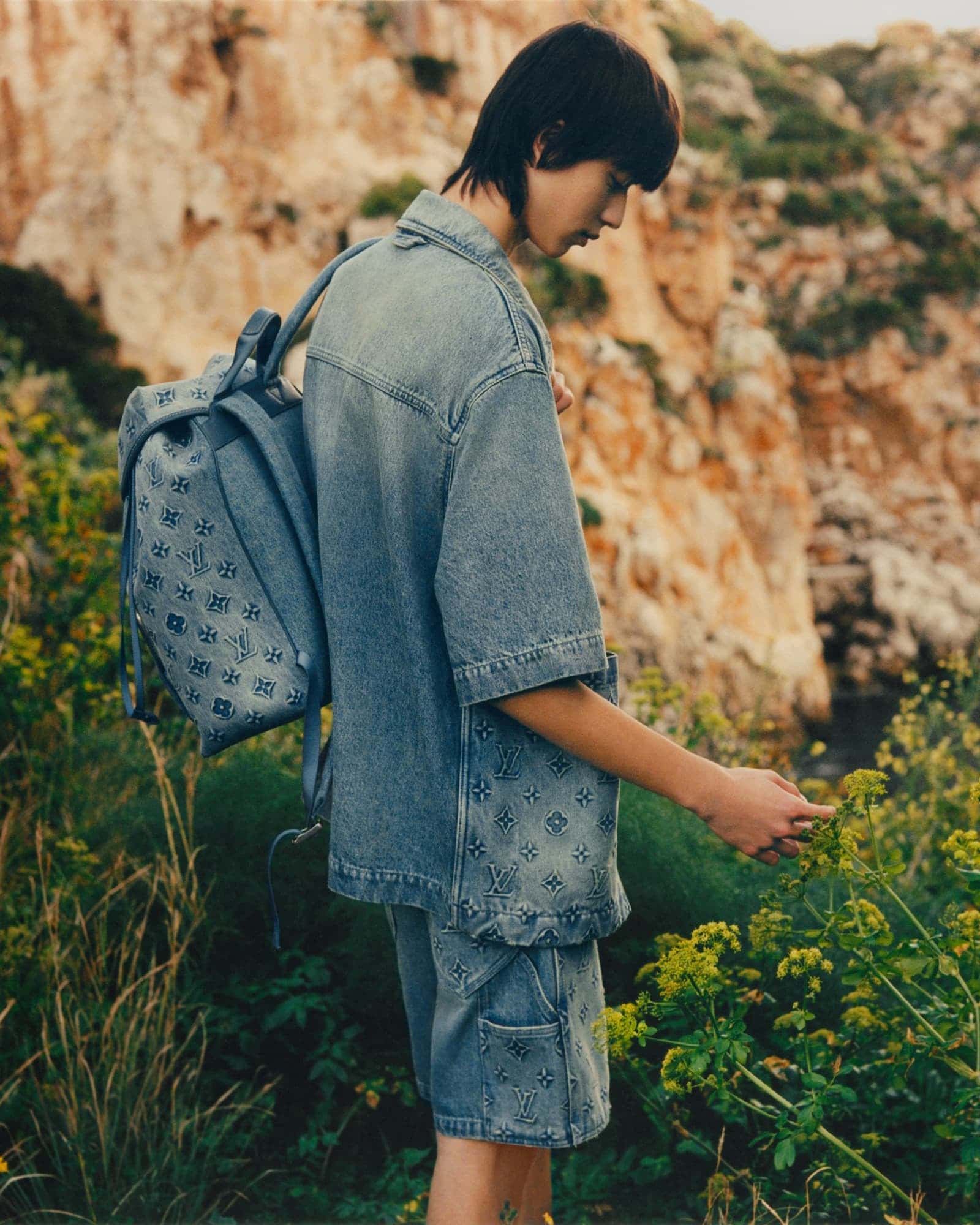 Man in denim shirt and shorts with a backpack, stands in a field with rocky hills in the background, side view, soft light.
