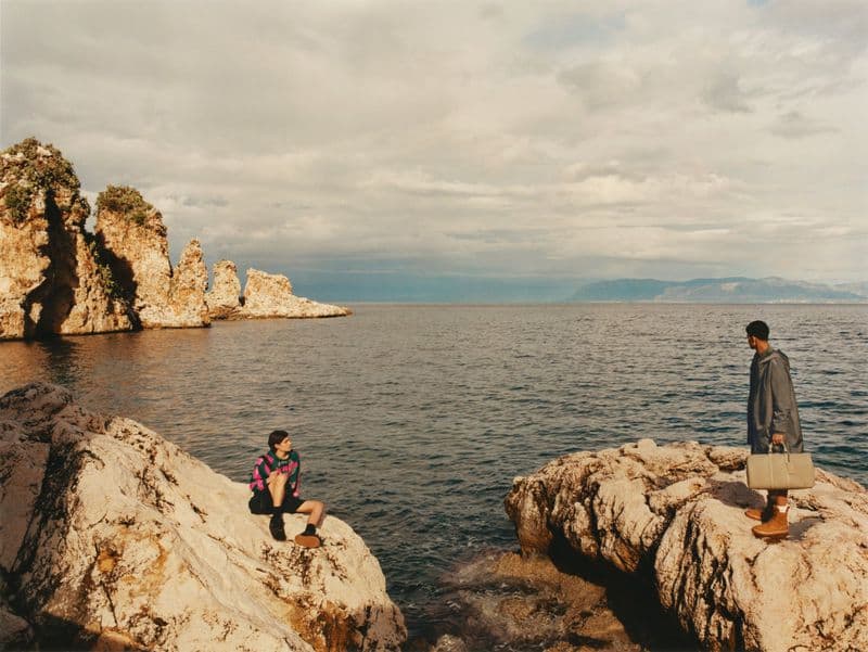 Two people on rocky coast near the ocean. One person sits looking out over the water, the other holds a briefcase near the shore.