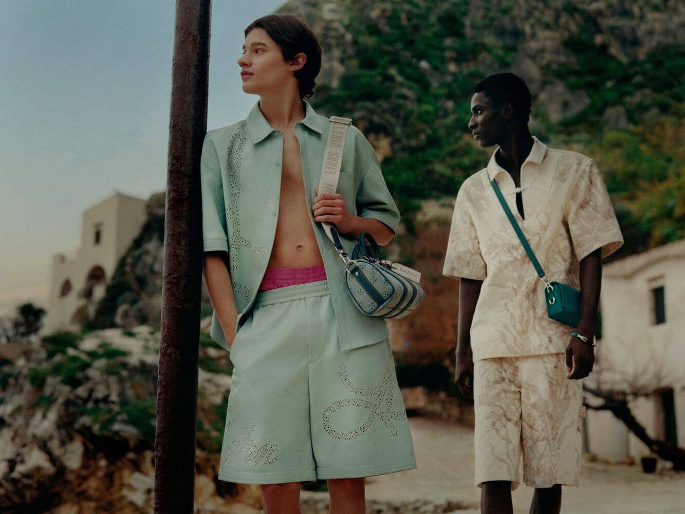 Two stylish men pose outdoors in matching patterned short sets and crossbody bags, set against a backdrop of mountains and buildings.