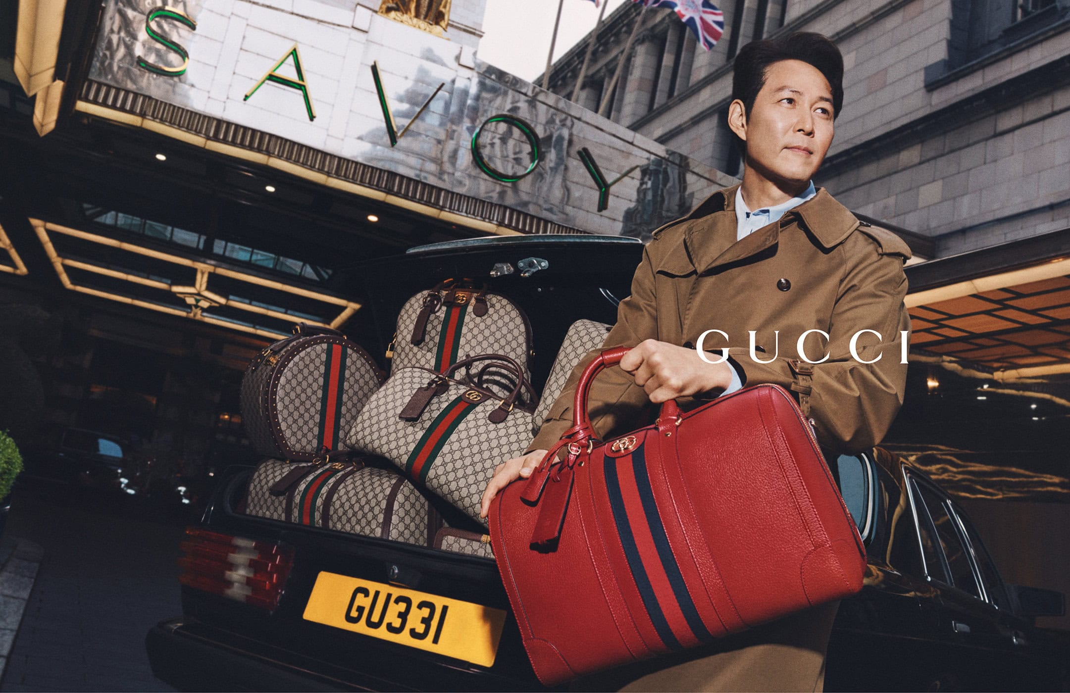 Asian adult male in trench coat holding red leather bag with luggage in car trunk in front of The Savoy Hotel for luxury brand ad campaign.
