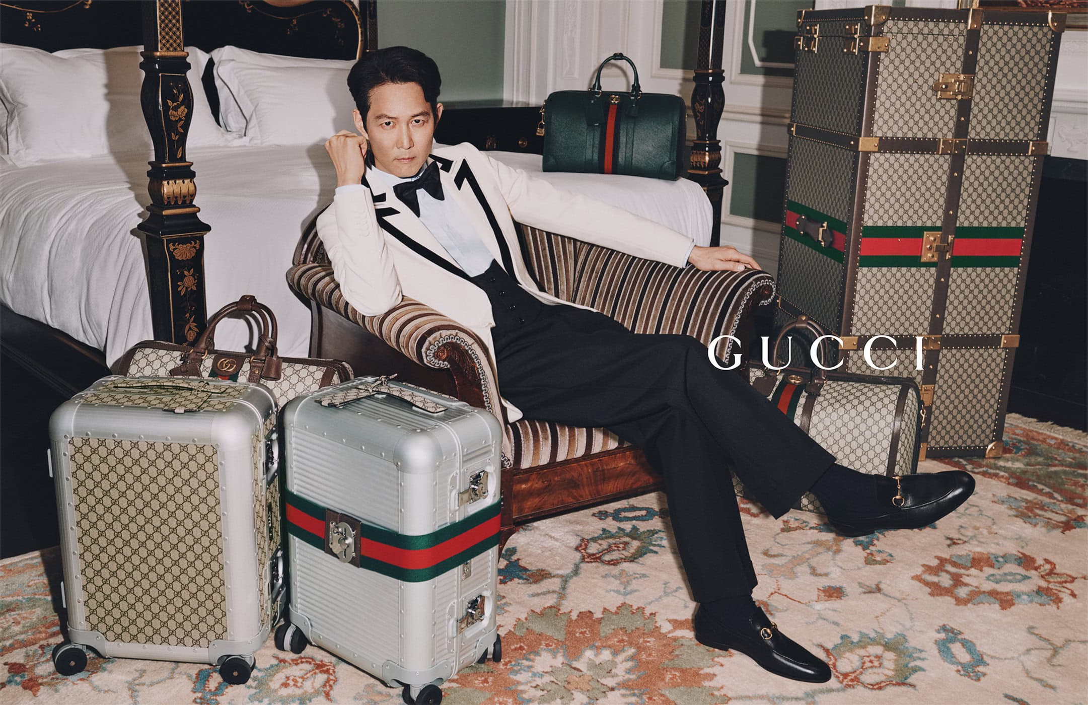 A man in a cream tuxedo poses in a vintage chair surrounded by Gucci luggage in a styled indoor hotel or apartment setting.