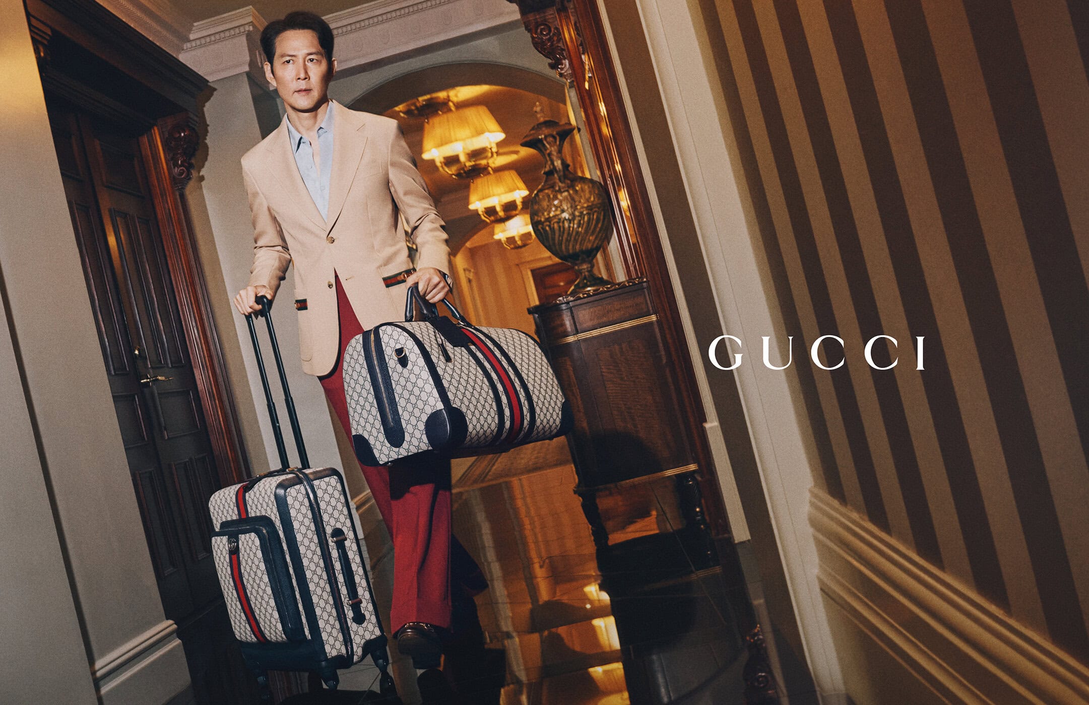 Asian man in a beige suit carries branded luggage in a hotel corridor with striped wallpaper for a fashion or travel advertisement.