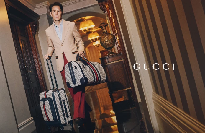 Asian man in a beige suit carries branded luggage in a hotel corridor with striped wallpaper for a fashion or travel advertisement.