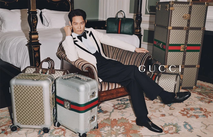 Actor Jungjae Lee seated on a striped armchair surrounded by Gucci luggage and trunks in a luxury hotel room setting.