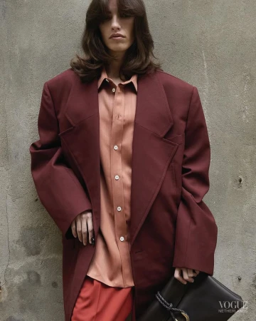Woman with long brown hair poses in a maroon oversized jacket and peach-colored shirt in front of a weathered concrete wall.