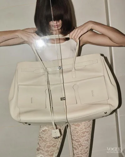 Model holding a large cream leather tote bag wearing a white top and lace trousers against a tiled wall