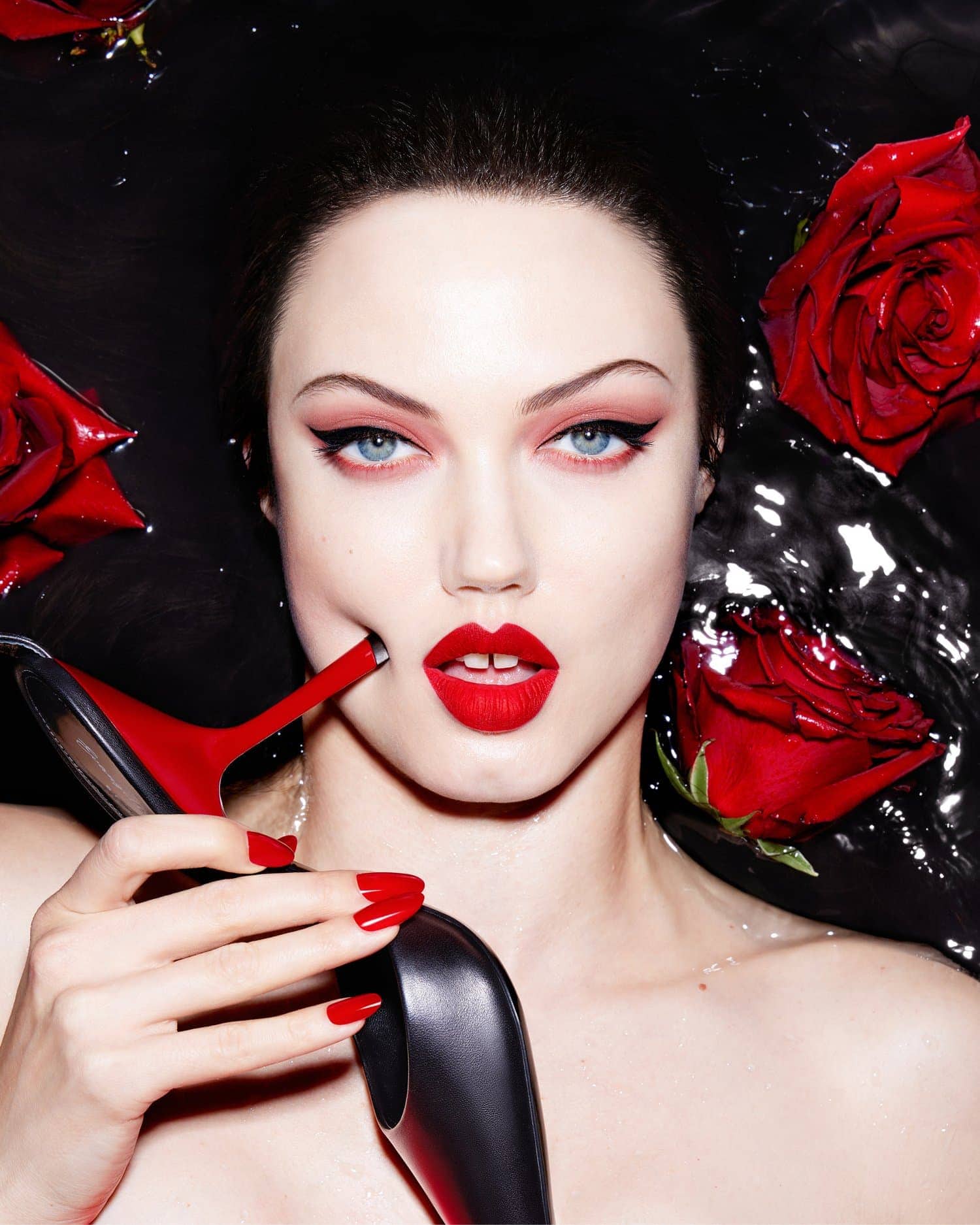 Beauty shot of a woman in the water with red roses, red lipstick and nails, holding a high-heeled shoe, dark backdrop.