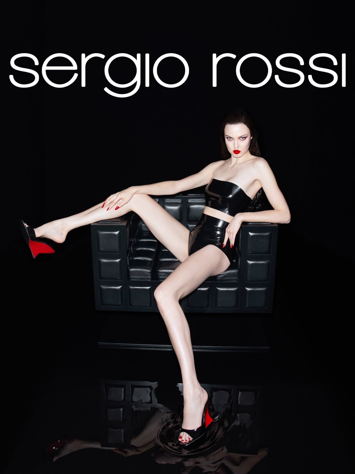 Woman poses in black patent leather crop top, shorts, and high heels sitting in a black chair against a black background.