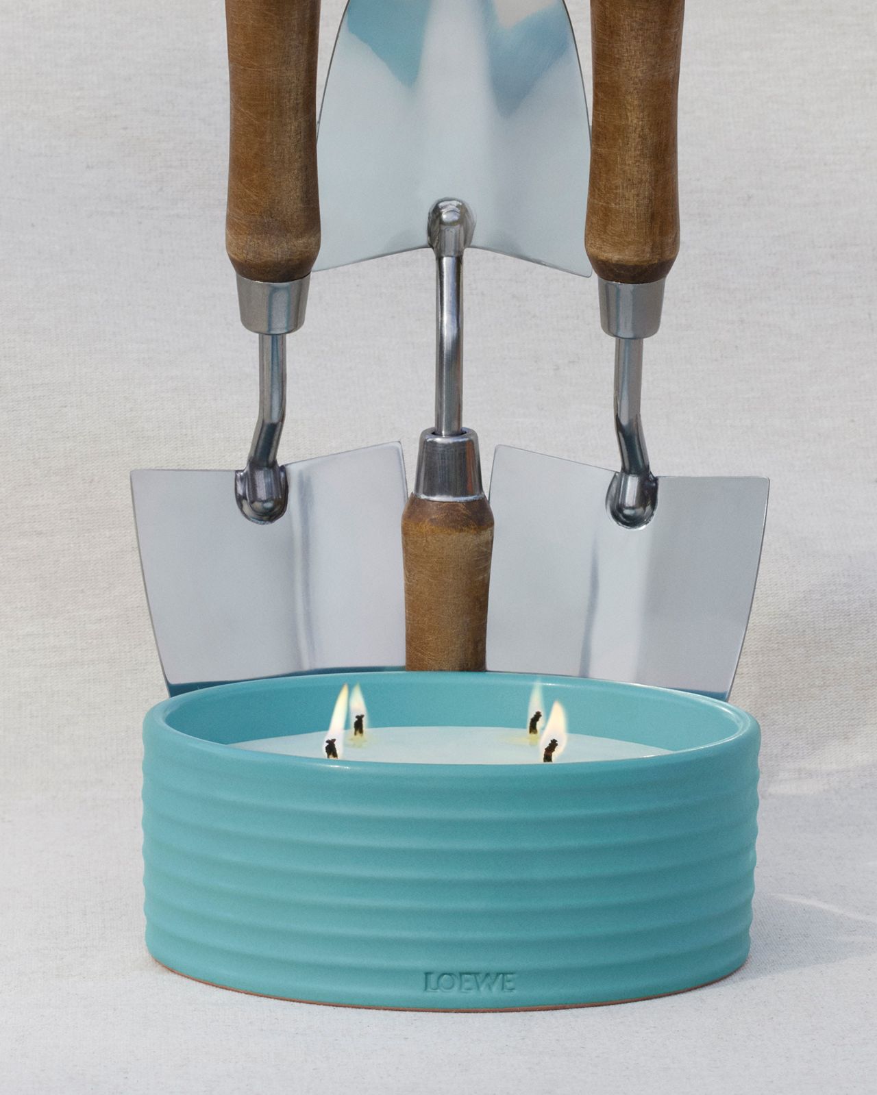 Still life of Loewe candle in a ribbed blue ceramic holder with lit wicks, backed by three gardening trowels on a light background.