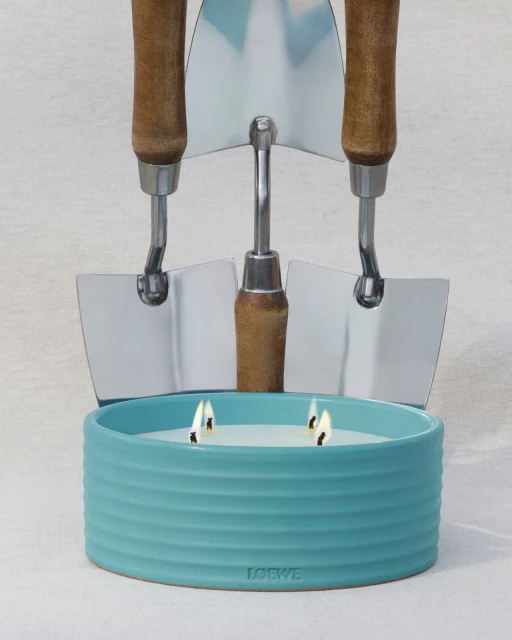 Still life of Loewe candle in a ribbed blue ceramic holder with lit wicks, backed by three gardening trowels on a light background.