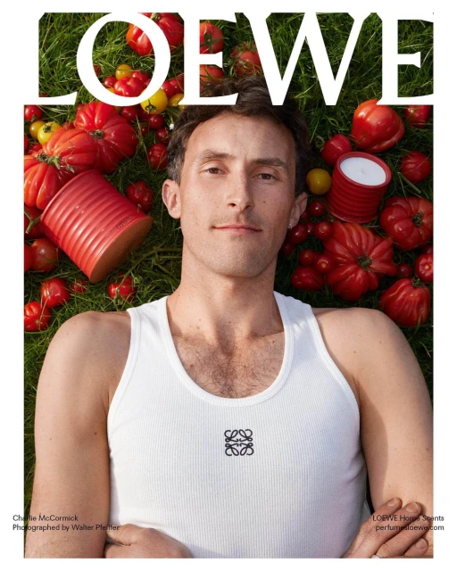 Man lying on grass wearing a tank top with tomatoes and red cans. The word 'LOEWE' is prominently displayed at the top.