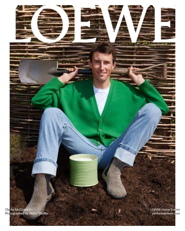 Male model in a green cardigan poses with a spade in a garden setting for Loewe Home Scents advertisement, showcasing natural textures and colors.