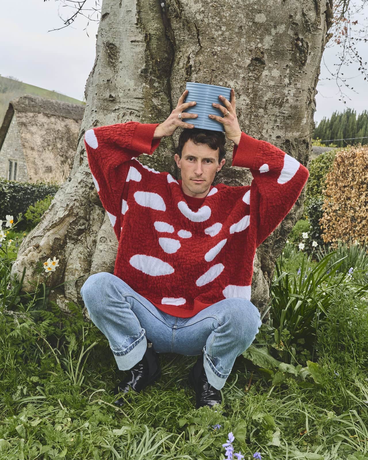 Adult male squats in a grassy outdoor area wearing a red mushroom-patterned sweater while holding a blue container on his head.