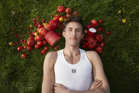 Man in white tank top lying on grass surrounded by tomatoes and candles for Loewe campaign