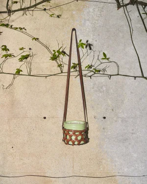 Loewe hanging scented candle in a light green ceramic vessel and woven brown leather basket against a textured wall.