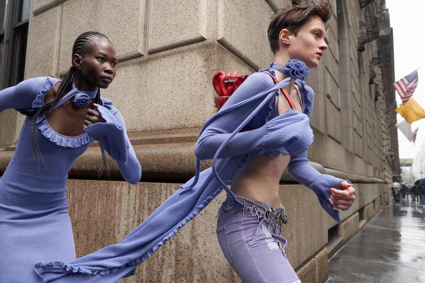 Two fashion models walking on a wet city street wearing matching periwinkle knitwear ensembles, with a vintage red bag on one's back.