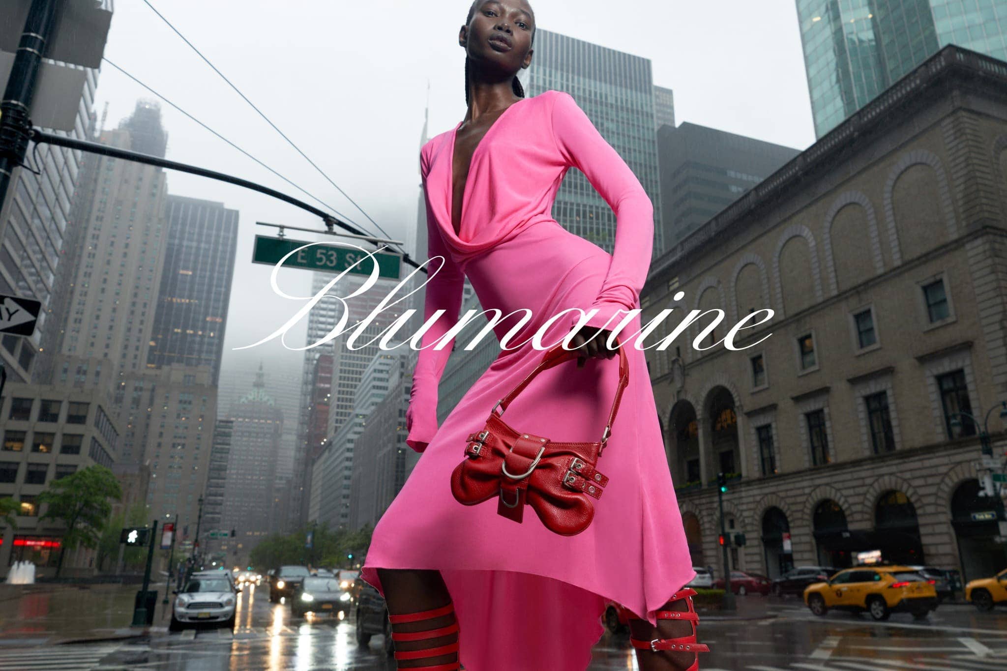 A Black woman in a pink dress and matching strappy heels walks on a city street carrying a red handbag, surrounded by urban buildings.