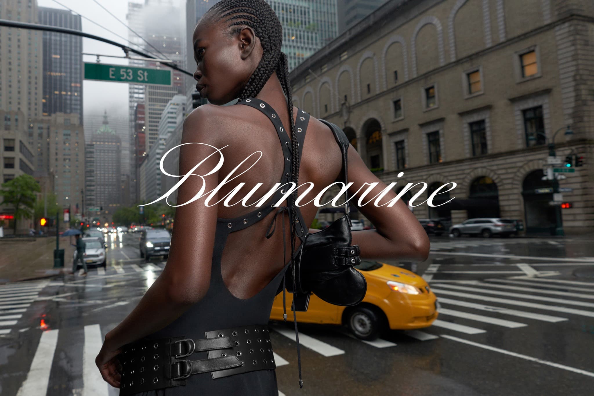 Fashion shot of African-descent woman in dress with braided hair, standing on city street with traffic and tall buildings in the background.