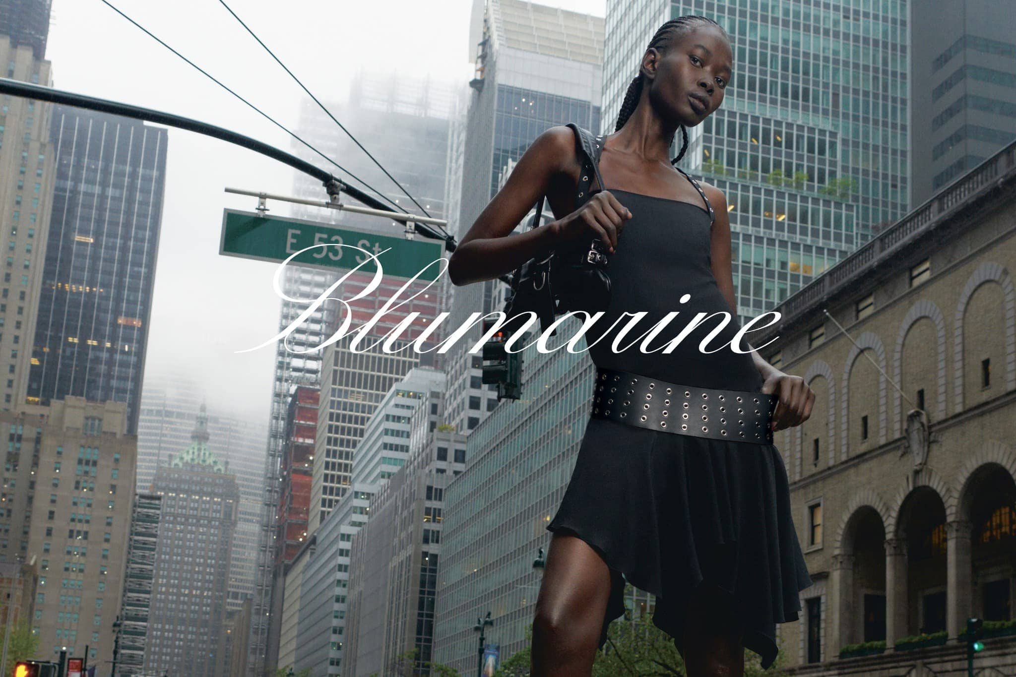 A woman in a black dress poses in an urban setting with skyscrapers. 'Blumarine' text overlay. Fashion editorial with city backdrop.