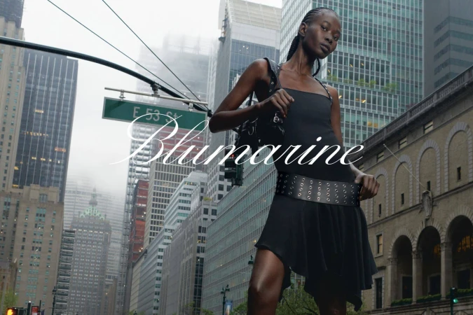 A woman in a black dress poses in an urban setting with skyscrapers. 'Blumarine' text overlay. Fashion editorial with city backdrop.