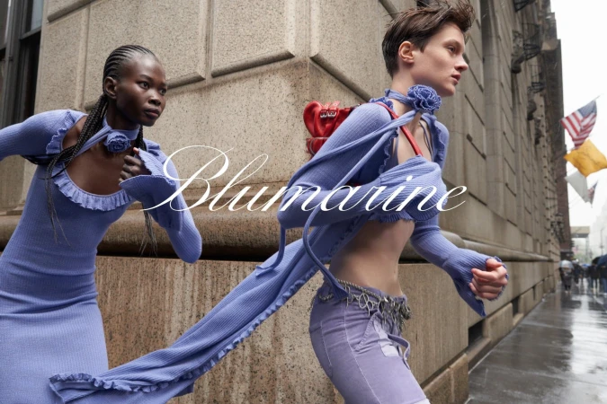Two models in lavender ribbed knitwear running past a stone building exterior with American flags