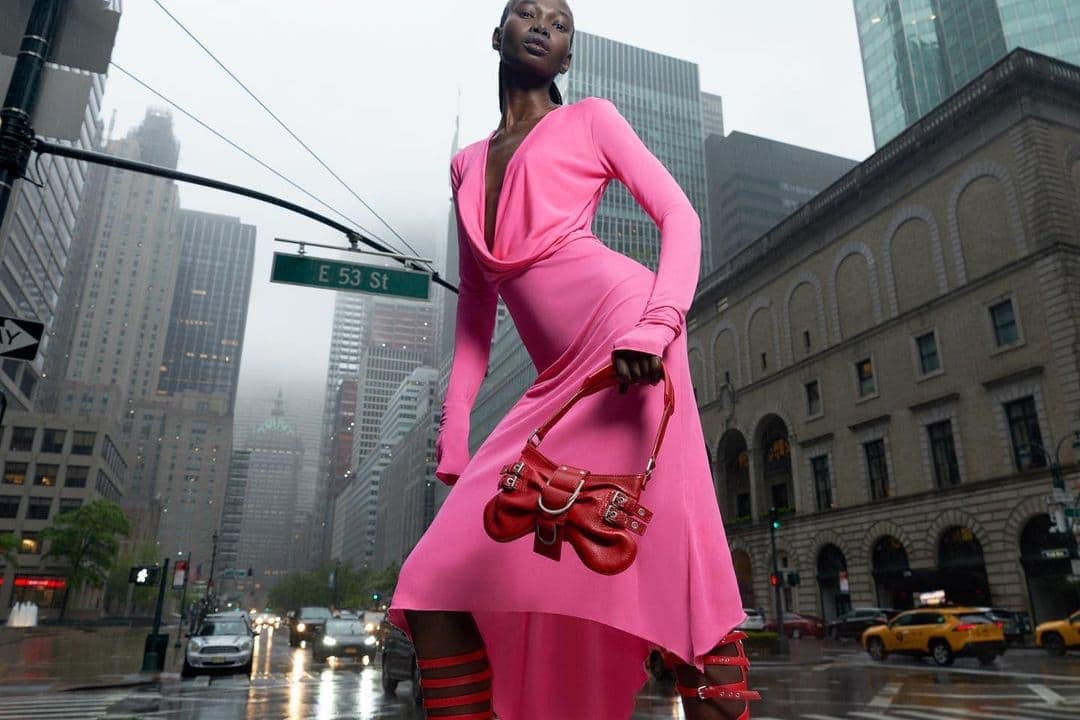 An African American fashion model poses in a pink dress with a red purse and gladiator-style heels on a rainy city street with tall buildings.