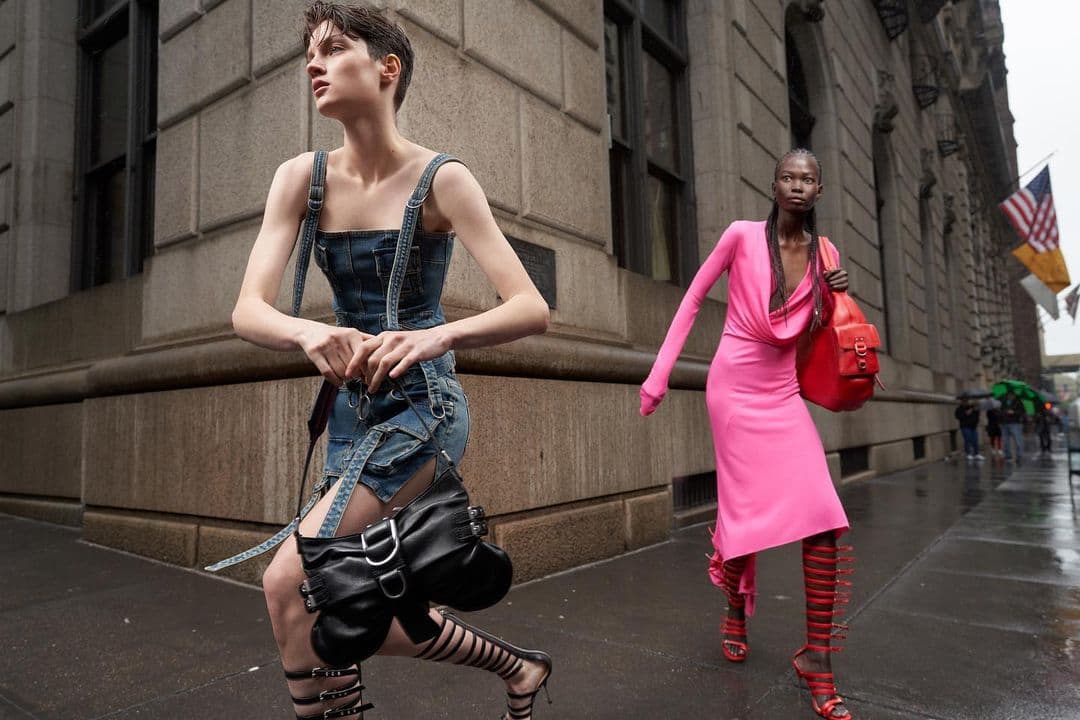 Two fashion models walk outdoors on a wet city street. One model wears a denim mini dress, the other wears a pink dress with a red purse.