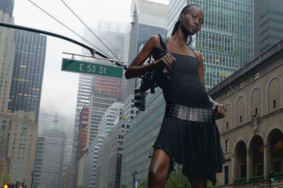Fashion shot of a Black woman in black dress with a wide belt posing on a city street with buildings in the background on an overcast day.