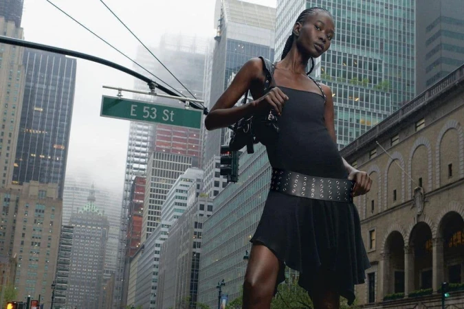 Fashion shot of a Black woman in black dress with a wide belt posing on a city street with buildings in the background on an overcast day.