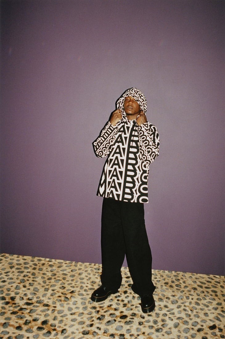 A person stands in front of a purple wall wearing a hooded jacket with a repeating logo pattern, paired with black pants and shoes.