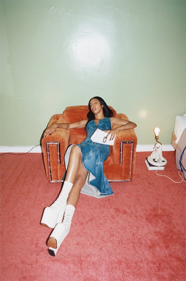 A woman in a denim dress poses in a vintage chair indoors, holding a white purse, with a lamp and books on a table nearby.
