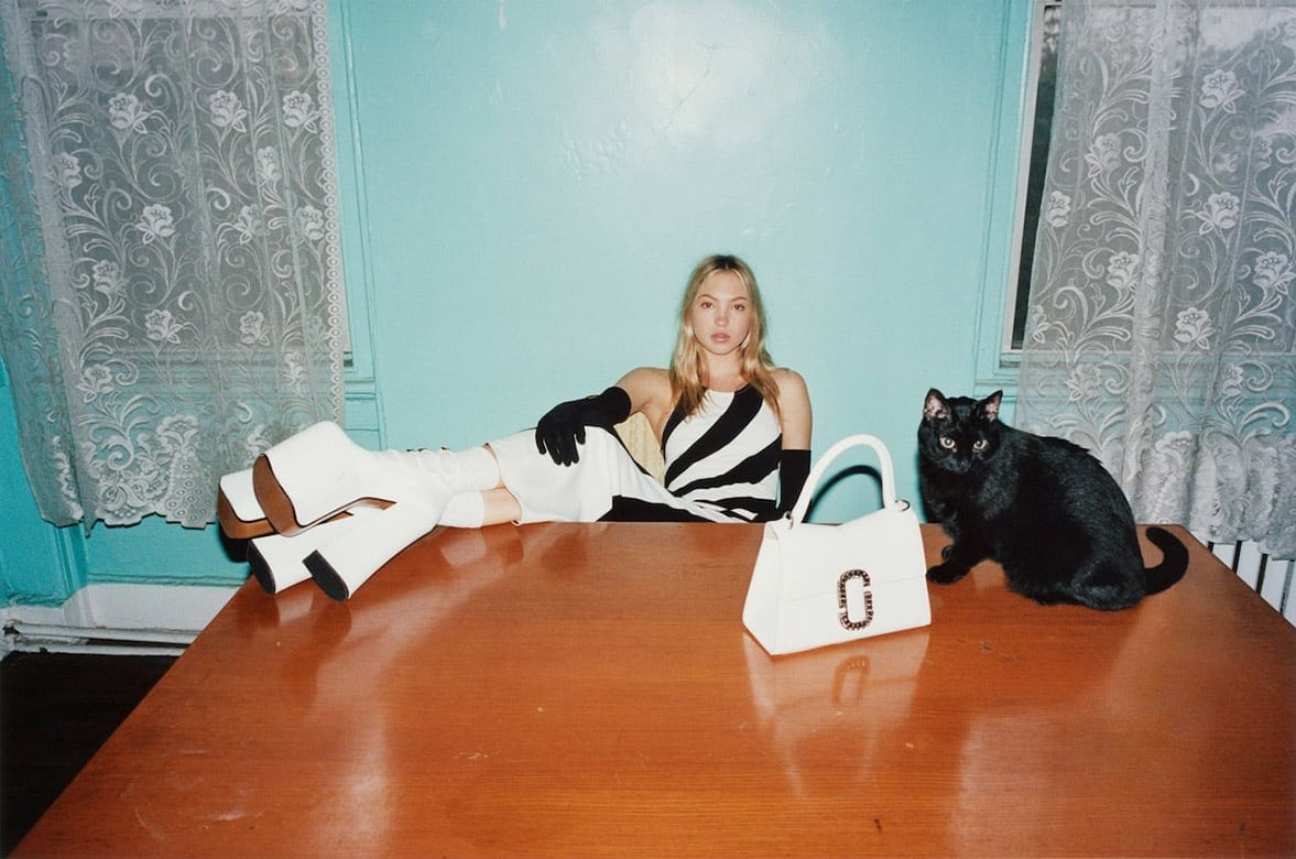 A woman poses on a table in a striped dress, white boots, and opera gloves, next to a white handbag and black cat against a blue wall.