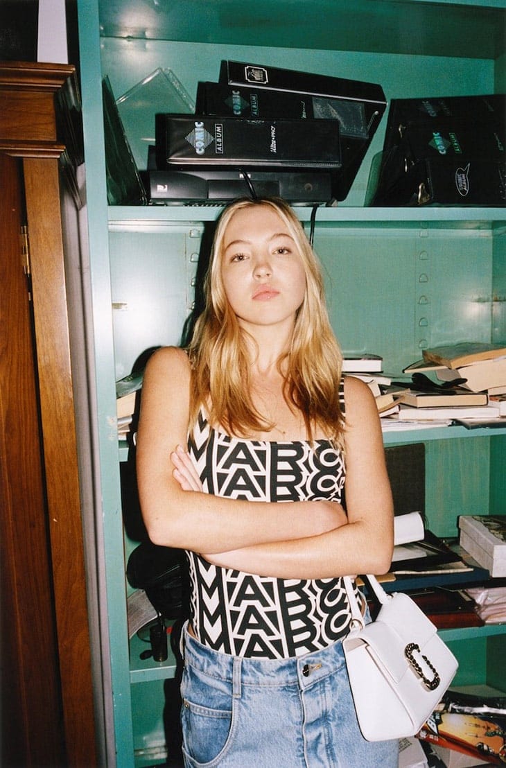 A blonde woman poses indoors wearing a Marc Jacobs print top, blue jeans, and a white purse, with a bookshelf in the background.