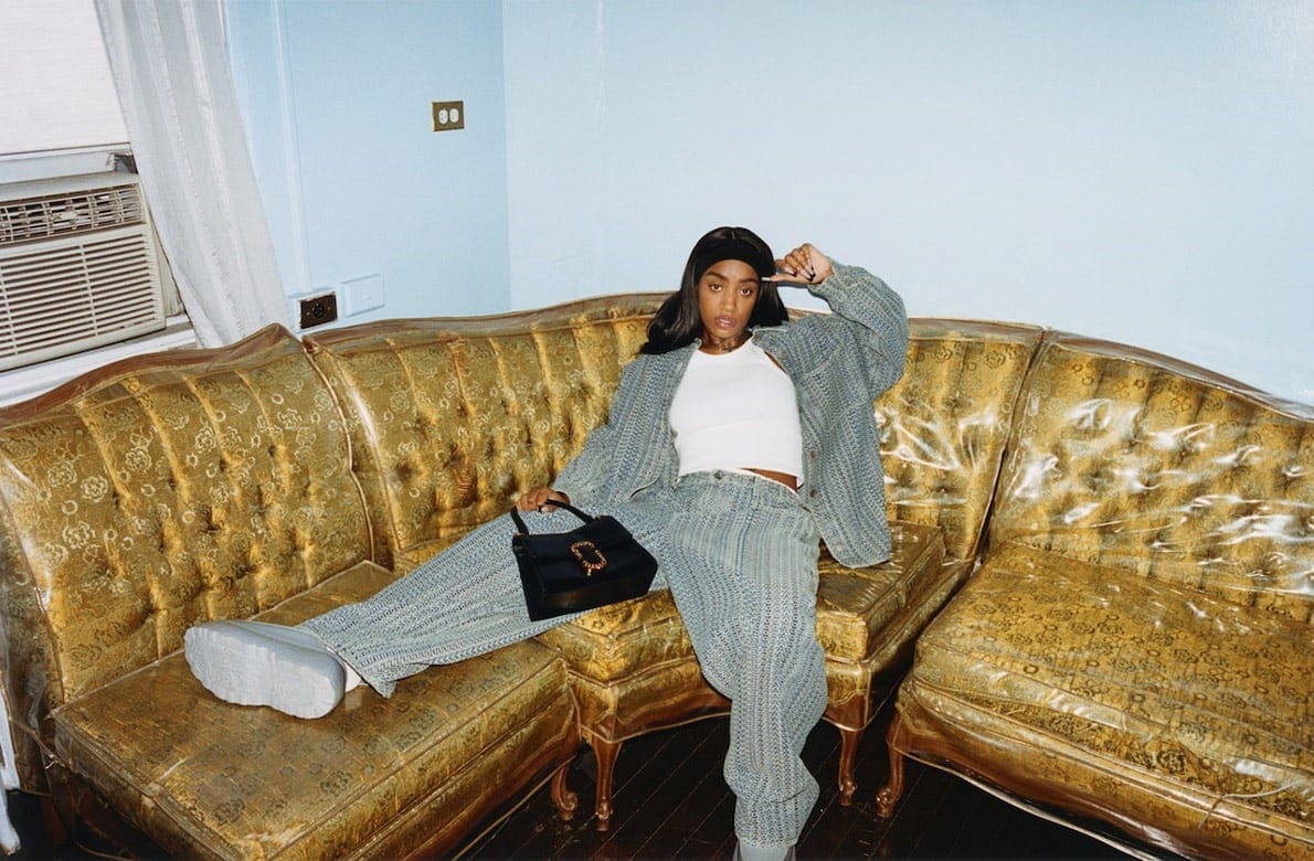 Woman in blue knit pantsuit poses on gold tufted couch in indoor setting with black purse, white top, and silver platform shoes.