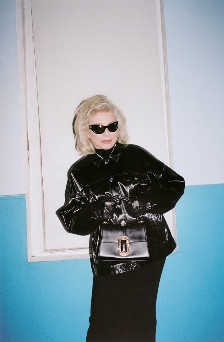 Elegant woman with platinum blonde hair poses in a shiny black jacket, black top and skirt with matching handbag against a two-tone blue wall.