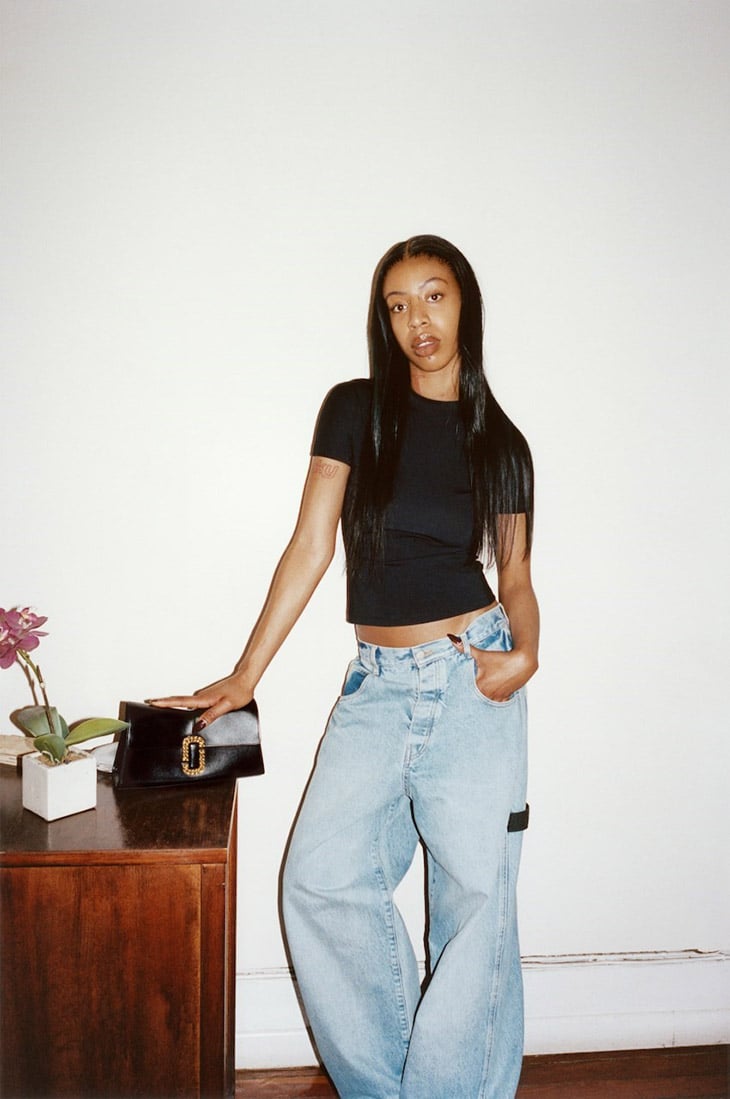 A young woman poses indoors wearing a black top and light blue baggy jeans, standing next to a dark wood desk with a purse and plant.