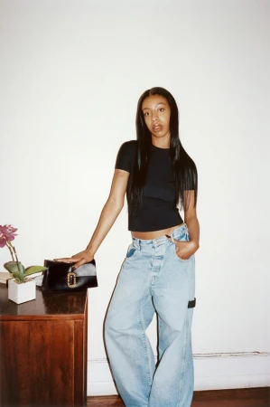 A young woman poses indoors wearing a black top and light blue baggy jeans, standing next to a dark wood desk with a purse and plant.