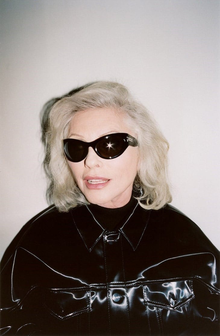 Blondie singer Debbie Harry in a studio portrait wearing sunglasses and a glossy black jacket against a gradient gray background.