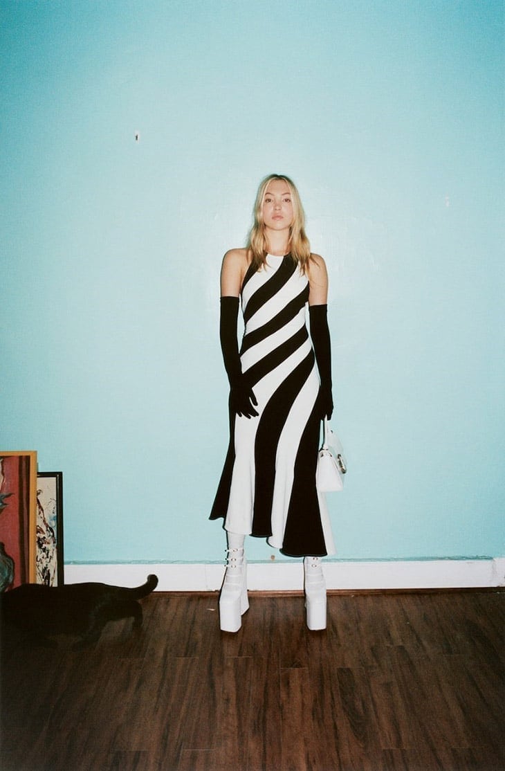 Fashion photo of a woman in a black and white dress, gloves, and platform boots, posing in a room with a black cat and art prints.