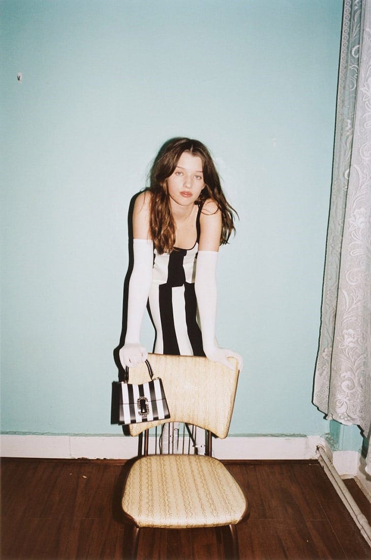 Young woman in a black and white dress leans on a vintage chair, holding a striped purse, posing against a light blue interior wall.