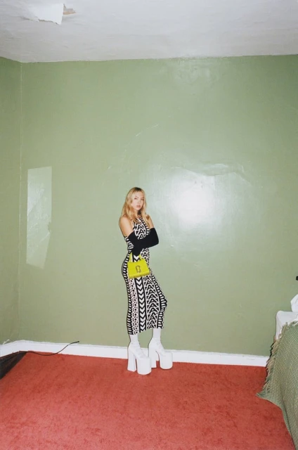 Woman poses in patterned dress with platform boots and handbag in a room with green walls and a red carpet.