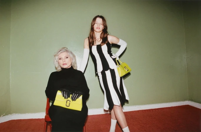 Debbie Harry seated in black holding a yellow clutch beside Ever Anderson in a striped dress holding a yellow handbag.