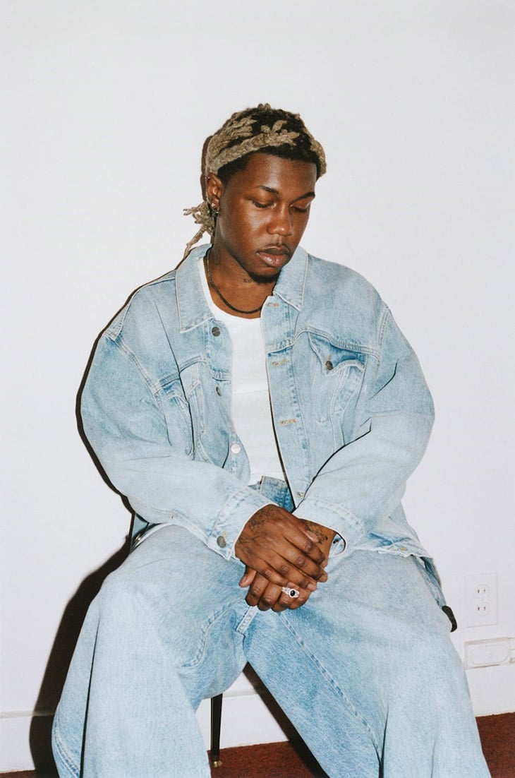 Portrait of a young adult male with medium brown skin in a light-blue denim jacket and pants, seated in an indoor studio setting.